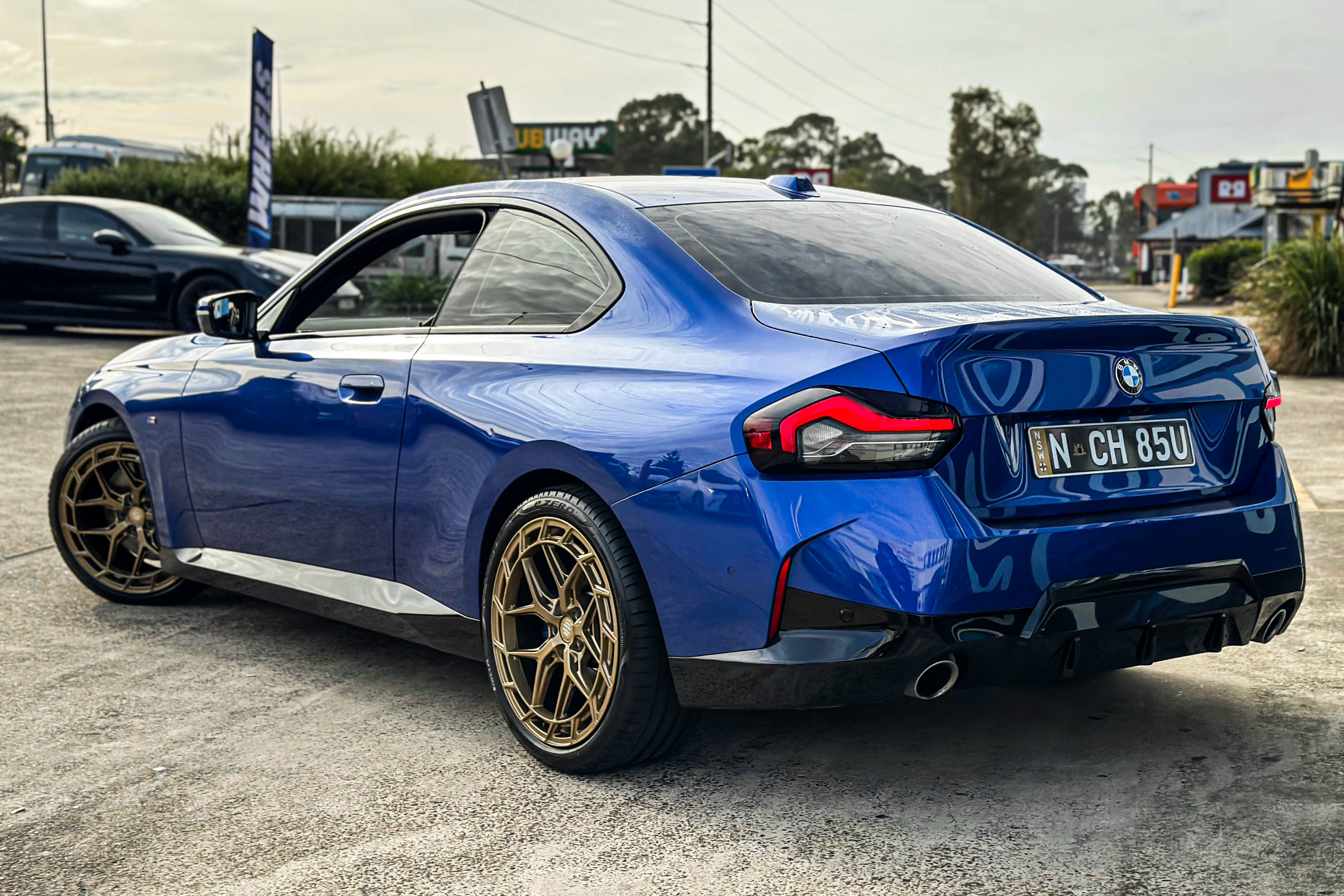 A blue BMW coupe with bronze multi-spoke wheels, viewed from the rear quarter
